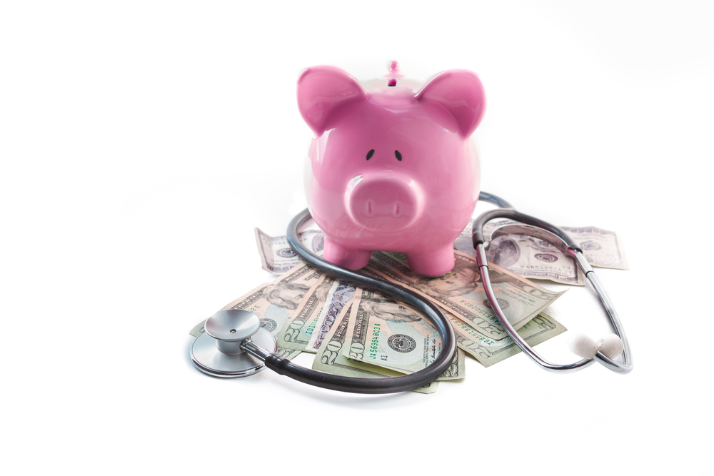 Piggy bank and stethoscope resting on pile of dollars on white background.